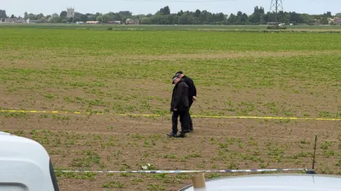 Two police officers walking in a field and peering down at the ground. The area they are in has been taped off with police tape. In the distance, are some buildings and a church steeple.