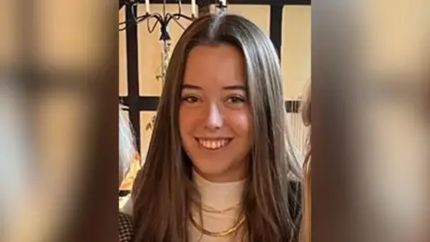 Image of sixteen-year-old girl with long brown hair smiling she is wearing a white high neck jumper with a gold chain over the top and is stood in front of a white wall with dark wooden panelling with a metal candelabra overhead.