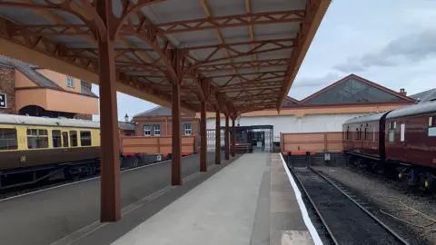 A drawing of a railway platform with old fashioned carriages on either side and a brown metal canopy above