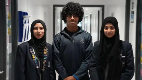 Three pupils stand side by side in a school corridor. Two are wearing dark blazers with a school badge, while the pupil in the centre is wearing a dark sports-style quarter-zip top.