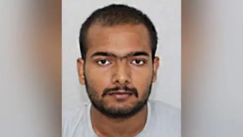 Custody photo of Pankaj Lamba against a grey background. He has dark facial hair, brown eyes and is looking at the camera.