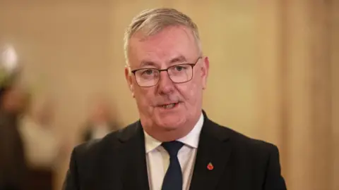 Nesbitt is looking at the camera and is speaking. He is wearing thin black framed glasses and wearing a black suit and tie. He has short grey hair. 