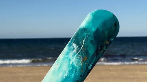 A picture of one of the painted body boards in Cornwall. It is mainly light blue in colour with the beach and sea in the background.