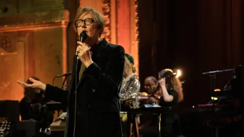 Pulp lead singer Jarvis Cocker performs on Later With Jules Holland while dressed in a brown velvet jacket and brown tie. He has wavy brown hair and a thick-rimmed pair of dark glasses.
