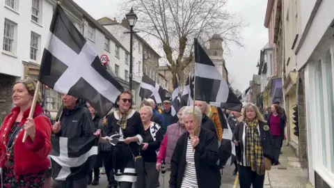 A picture of a number of people walking through Penryn with flags. There is shops either Side of the road.