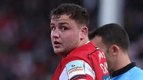 Gloucester prop Ciaran Knight in a cherry and white shirt looks towards the camera, with a blue-shirted referee standing just behind him.