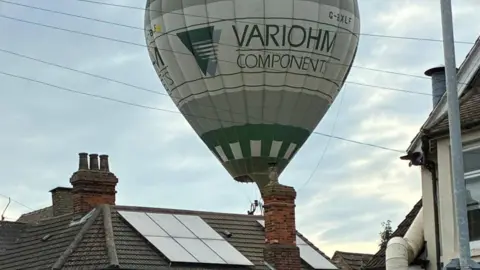 Ian Cording A hot air balloon hovering over roofs in Bedford. The balloon is green and grey and you can see solar panels, chimneys, poles and wires.