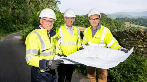 Three people wearing white hard hats, safety glasses, and high-visibility yellow jackets stand beside a rural road, holding and reviewing a large site plan. The tree men are all all clean shaved and smiling. The man on the right in Tim Farron. Trees and rolling green hills are in the background. The sky is blue. 