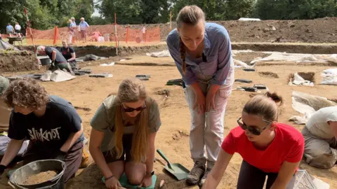 A woman in a blue shirt leabs over students, including a young woman in a red t-shirt and sunglasses, working in front of a series of excavated gaves.