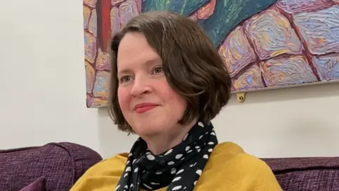 A white woman with brown hair and brown eyes, wears a black spotty scarf, yellow jumper and black leggings. She sits on a purple sofa in front of a colourful art canvas.