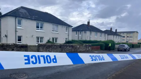 blue and white police tape which is upside down in front of a row of cream house. A police officer is visible in a distance.