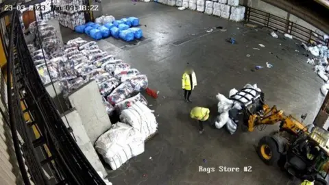 HSE A wide shot from a CCTV camera looking down onto a warehouse floor.
There are a large number of bundles of clothing. In the foreground, two people in high-vis jackets can be seen near to the front of a yellow machine with prongs on the front holding white textiles