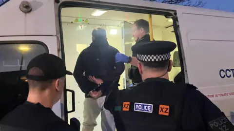 Photograph of a man being taken into custody after he was arrested during police raids in Mossley. The image shows a suspect being placed into the back of a police van.
