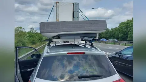 A grey car which is parked on the side of the M11 motorway with the door open. Attached to the roof of the vehicle is an outdoor foldable table with a mattress on top and then a white fridge on the top of the pile.