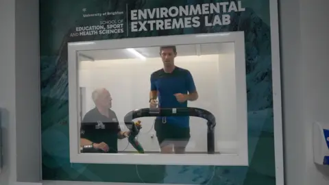 A man running on a treadmill behind a glass window. Another man is stood next to him.