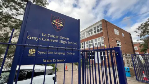 Stuart Woodward/BBC The entrance to a school, with a navy blue sign behind blue iron railings. There is a three-storey brick building behind it with white windows.