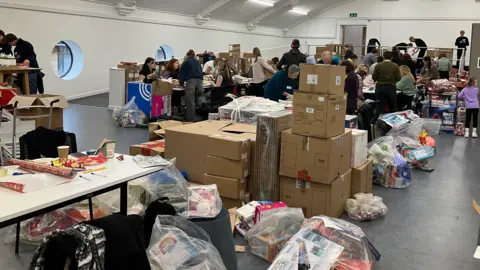 A large room is pictured with many boxes and bags of gifts visible on the bare floor. Dozens of people are also visible packing presents and talking to each other. The walls are white and have porthole-style windows