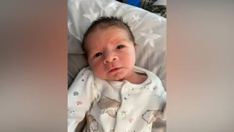 A small baby with short, dark hair is lying on a grey pillow/bedding set with stars on. He is wearing a white babygrow with a bear pattern on it.