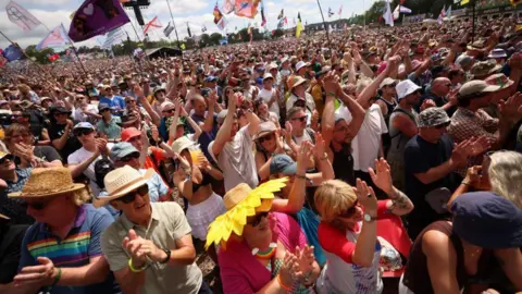 Festival-goers, many wearing hats and sunglasses, clap during a set at Glastonbury as the sun beams down on Friday