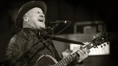 Karen Miller/BBC Black and white photo of a man with a goatee beard wearing a hat, singing and playing a guitar
