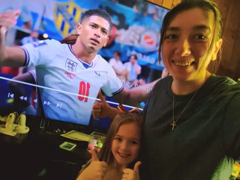 Courtney Silbaugh A woman and a child give the thumbs up while taking a selfie together. Behind them is a television showing England footballer Jude Bellingham in a white jersey with his hands in the air.