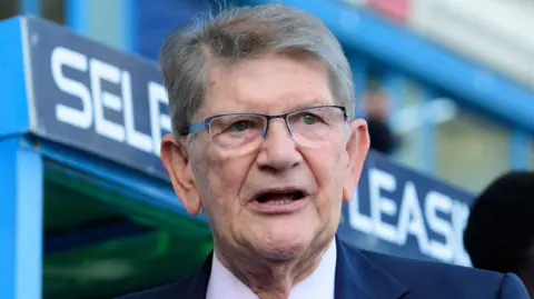 A picture of Sir John Madejski, who has greying hair and glasses and is wearing a suit, in front of a dug out at the SCL Stadium in Reading.