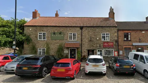 Google A Google Maps image of the Hansom Restaurant in Bedale, a large stone building that neighbours a pizza takeaway. The building has five large windows with white frames, and a black drain pipe down the middle dividing the two properties and their front doors. Hansom has a tiled awning above their black door, and there are seven cars parked along the street in front of it. The property has a chimney, a tree growing directly next to the restaurant, and greenery is growing up the bricks. There is also a street light off to the left.