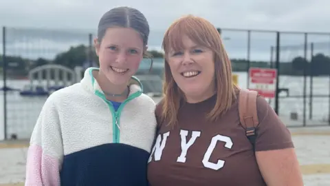 A mother and daughter are standing together outside in front of a black fence. They are both smiling at the camera. Behind the fence is a large inflatable water park.
