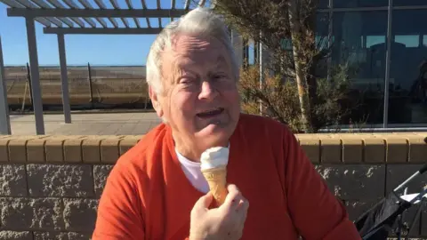 Supplied Bill Trotter is a older man in his seventies who is wearing a red jumper and a white teeshirt. He has grey hair and is eating an ice-cream. He is sitting in front of a wall and just behind him you can see a pram