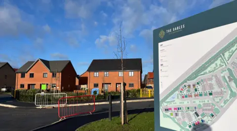 Shaun Whitmore/BBC A road into a housing development curves away to the right. To the right of the picture a sign says "The Gables Attleborough" and shows a map of the housing development. Straight ahead are two red brick houses, with a small green space and a play park in front of them.