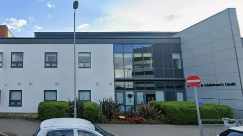 Google External view of Blackpool Victoria Hospital's Women's and Children's Unit in Blackpool, Lancashire. The image is taken on a sunny day. Cars are parked outside the building.