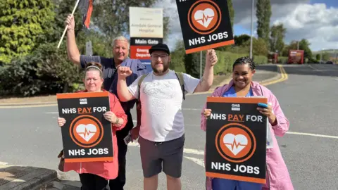 Aisha Iqbal/BBC A group of four people stand on a roadside holding placards and flags saying "NHS Pay for NHS Jobs". One of them wears hospital scrubs. Behind them is the entrance and sign for Airedale General Hospital.