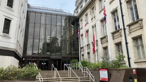 Lewis Adams/BBC County Hall in Chelmsford. It has a glass facade and has four flagpoles out the front. Stairs lead to the entrance.