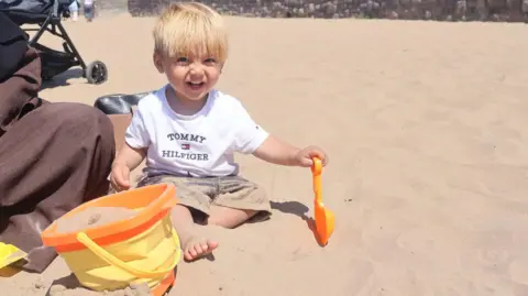 A young boy with short blonde hair sits in a white t-shirt and dark shorts on the beach. He is smiling and holding an orange spade, covered in sand. Next to him is an orange and yellow bucket full of sand. 