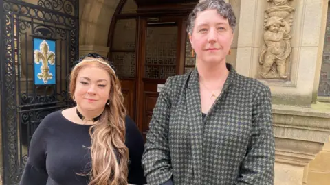 Two women stood outside a council building. The woman on he left has long blonde hair with a headband and is wearing a black top and black choker-style necklace. The woman on the fight has short, dark grey hair and is wearing a grey and black checked jacket. 