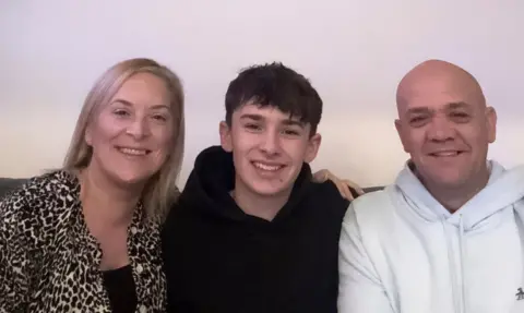Finley with mum Kelly and dad Mark. Kelly has long blonde hair and wears a black top with leopard print jacket. Finley has a black hoodie on. Mark is bald and wears a white hoodie.