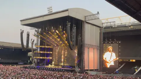 Ed Sheeran performs on stage while playing a guitar. Thousands of people watch from below and in front of the stage.