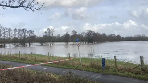 Dorset Council A rural area in Dorset with severe flooding. A field has almost completely been submerged under water. A white and Red ribbon to prevent people from going further up the path beside it has been put up.