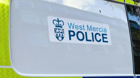 A West Mercia Police sticker on a police van which is predominately white on the panel pictured with yellow high viz stickers around it.