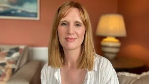 A woman with red hair and white shirt. She is sitting in a living room with a lamp in the background and peach coloured walls. There are beige sofas and a painting on the wall.