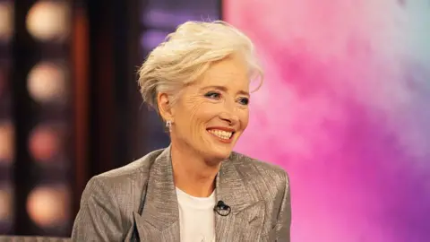Emma Thompson sat on stage on the Kelly Clarkson show. She is smiling whilst wearing a white t-shirt and silver blazer. 