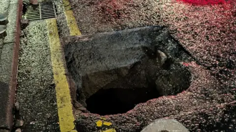 A close-up of the sunken manhole on Bridge Street, Newry at night. It has left a gapping hole in the road surface. There are double yellow lines running along the side of the pavement, but part of the lines are missing as the manhole cover has disappeared from view. 
