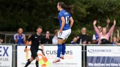 Ross Halls Sophie Peskett, jumping in celebration, wearing the blue Ipswich Town it, as fans raise their arms in the background. 