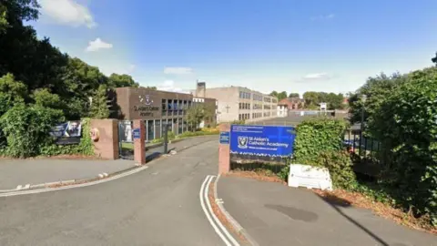 Google St Aidan's Catholic Academy. Two brown brick school buildings can be seen inside the grounds. A religious cross is displayed on one wall. There are trees and plants growing around the school's border.