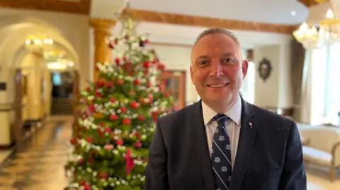 Andrew is smiling at the camera. He has short dark brown hair and is clean shaven. He is wearing a white shirt with a royal blue tie and suit jacket.