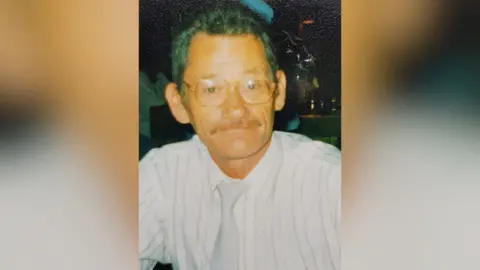 Eileen Burkert A man in his fifties, with short brown hair and glasses is looking at the camera. He has a moustache and is smiling. It is an old photograph and some of the colour has drained from it. He is wearing a shirt and tie.