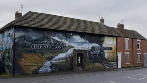 Disused pub The Station, in St Helen Auckland, and two adjoining two-storey brown brick houses with boarded up windows. The pub building has a painting of a black steam train going through a mountain landscape. There is a writing reading The Station Free House. A viaduct is painted on the side wall of the pub. There are gins and notices on its black door. 

