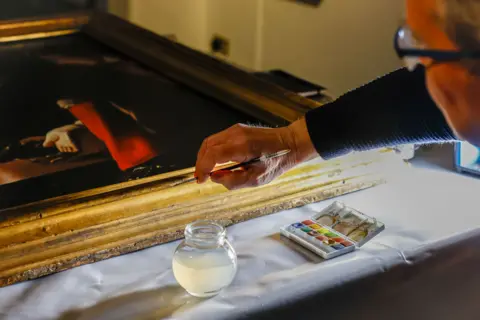 Stockton Borough Council A conservator holds a paintbrush to the frame of The Dice Players for restoration work. The face of the conservator is only partially in view, with the focus on his hand working on the golden-coloured frame.