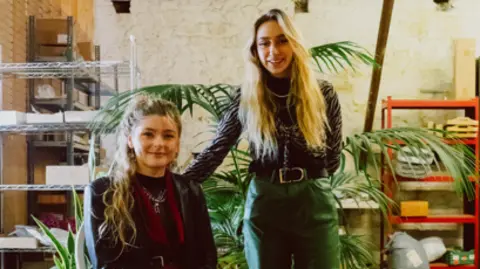 Sophie Harris is sat on a chair to the left and Hayley Gibaut is standing to the right. The pair are inside of the space. Green plants are behind them. Ms Harris is wearing dark clothing and has long light hair. Ms Gibaut is wearing dark clothes and has long blonde hair.