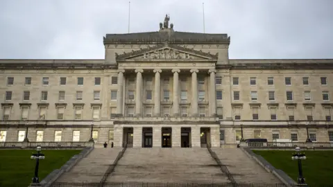 Liam McBurney/PA Wire A view of Parliament Buildings from beneath the steps of the Stormont Estate. It is a large, detached, four-storey over basement Neoclassical Portland limestone construction with six pillars on its central portico. The are neat lawns and two black cast-iron standard lamps on either side of the steps.  A statue of Britannia, flanked by two lions, is on top of the building.